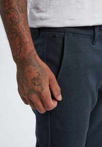 Navy textured trousers with a subtle checked pattern, featuring a front pocket and a metal hardware accent near the waistband.