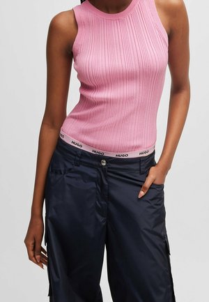 Femme portant un top rose sans manches en côtes et un pantalon bleu marine, une main dans la poche et l’autre détendue le long du corps.