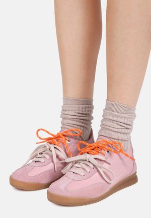 Beine mit pinken Sneakern mit beigen Sohlen, unterschiedlich beigen und knallorangen Schnürsenkeln und lässigen beigen Socken vor einem schlichten Hintergrund.