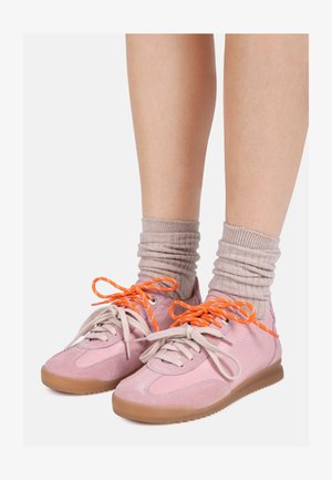 Benen met roze sneakers met beige zolen, niet-matching beige en feloranje veters en losse beige sokken tegen een effen achtergrond.