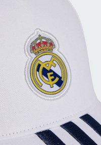 White cap featuring an embroidered Real Madrid logo in blue and yellow, crowned with red details, along with navy stripes on the brim.