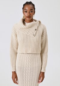 Maglione beige lavorato a maglia con collo a cowl, caratterizzato da un motivo diagonale a coste, tre bottoni argentati su una spalla e design cropped.