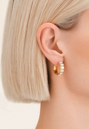 Boucles d'oreilles en métal doré en forme de créole, ornées de cristaux rectangulaires transparents, d'une surface texturée et d'une fermeture à charnière, portées sur une oreille avec des cheveux courts.