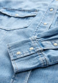 Lichtblauw denim overhemd met zilveren drukknoopjes, voorzien van een opgerolde manchette en twee borstzakken. Toont zichtbare stiksels en heeft een zachte textuur.