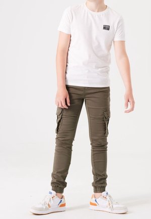 Kind trägt weißes kurzärmeliges Hemd und olivgrüne Cargohose mit elastischen Bündchen, steht mit leicht auseinander stehenden Füßen vor weißem Hintergrund.