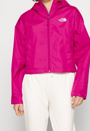 Vrouw draagt een felroze North Face jas met capuchon en rits en witte broek, getoond van nek tot heupen tegen een effen achtergrond.