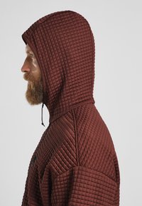 Braune gesteppte Kapuzenjacke mit großer Kapuze, strukturierten Details und Kordelzug. Der Stoff wirkt weich und dick und bietet Wärme.