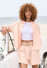 Peach linen oversized shirt, white ribbed crop top, matching peach shorts with a beige belted waist, against a beach and vintage car backdrop.