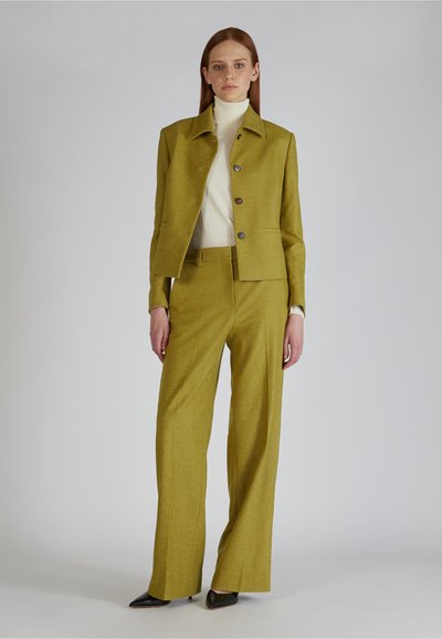 Vrouw staat in een olijfgroen tailored pak met een geknoopte blazer en brede broeken, witte coltrui en zwarte puntige schoenen.