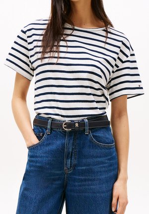 Kvinde iført hvid og navystribet kortærmet skjorte, stoppet ned i højtsiddende blå jeans med sort læderbælte med guldfarvet spænde.