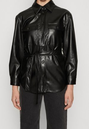 Chemise noire en similicuir à boutons, manches longues, poches poitrine et ceinture à la taille, portée avec un pantalon gris foncé.