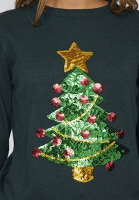 Mörkgrön tröja med en paljettdekorerad julgran design, med en gyllene stjärna på toppen, röda prydnader och en brun stam.