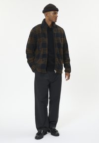 Giacca in pile con zip in un motivo a quadri blu navy e marrone, abbinata a un maglione nero e pantaloni neri larghi. Scarpe nere voluminose completano il look.