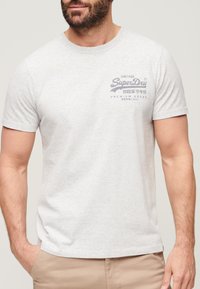 Lysegrå crew neck t-shirt med korte ærmer, der har et mørkeblåt grafisk tryk på brystet. Stoffet ser blødt og struktureret ud.