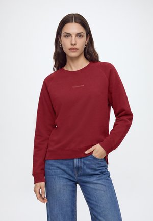Ung kvinde med langt brunt hår iført en rød sweatshirt og blå jeans, stående med den ene hånd i lommen mod en ensfarvet baggrund.