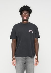 Sort bomulds t-shirt med rund hals og korte ærmer, prydet med et lyserødt "Tommy Jeans" logo buet over venstre bryst. Bæres med grå jeans.