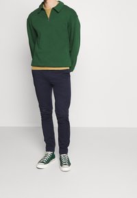 Grön zip-up huvtröja med krage, lager på lager över en beige skjorta, kombinerad med marinblå byxor och gröna sneakers.