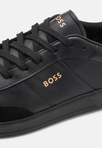 Svart lädersneaker med texturerade tygalternativ, med platta svarta snören och präglad "BOSS"-logga i guld på sidan.