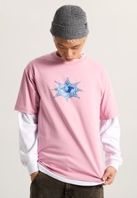 Camiseta de algodón rosa claro con un diseño gráfico de estrella azul en el centro. Llevada sobre una camisa de manga larga blanca, combinada con jeans oscuros.