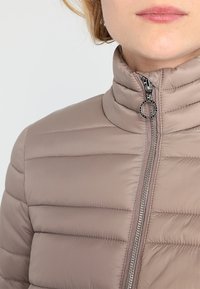 Licht taupe pufferjack met horizontale doorgestikte textuur, hoge kraag en metalen ritsdetail. Gladde stof met een zachte afwerking.