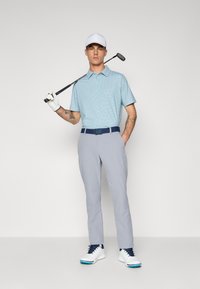 Golf polo shirt in light blue with a dotted pattern, gray pants, navy belt, and white golf shoes. The golfer holds a club and wears a white cap.