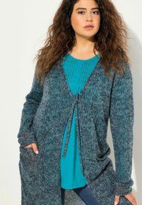 Virkat cardigan i mörkt teal med ett strukturerat stickat mönster. Har en knytning i midjan och två sidofickor. Lagd över en teal topp.