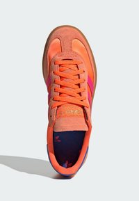 Orange suede sneaker with pink stripes, textured upper, and rubber sole. Gold logo on tongue and patterned insole with blue and red accents.