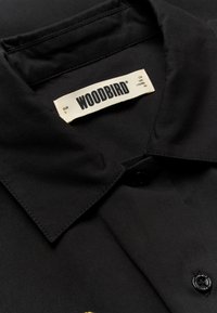 Chemise noire à col montrant une étiquette de marque WOODBIRD blanche cousue à l'intérieur de l'encolure avec les détails de la taille.
