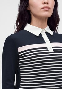 Femme portant un haut bleu marine avec des rayures horizontales blanches, une rayure rose clair et un col blanc boutonné, vue des épaules au menton.