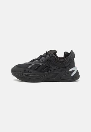 Baskets de sport noires avec semelle épaisse, tige en mesh et cuir, bandes latérales et un accent incurvé blanc au talon.