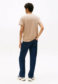 Mand med krøllet hår iført en beige t-shirt, mørkeblå jeans og hvide sneakers, stående med den ene hånd i baglommen mod en hvid baggrund.