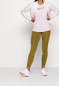 Jasnoróżowa bluzka z krótkim rękawem z kwiatowym logo, zestawiona z oliwkowymi legginsami i białymi butami sportowymi z różowymi akcentami. Stylowa kurtka przeciwdeszczowa.