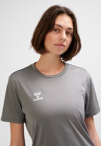 Sportowa koszulka z jasnoszarego materiału o gładkiej fakturze. Posiada krótkie rękawy i okrągły dekolt, ozdobiona białym logo na lewej piersi.