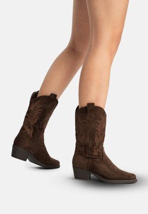 Jambes portant des bottes western en suède marron foncé mi-mollet avec des motifs brodés et des talons bloc bas, un pied légèrement levé.