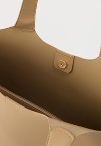 Τσάντα χειρός από μπεζ δέρμα με απαλή υφή, ανοιχτό πάνω μέρος και κουμπί κλεισίματος. Το εσωτερικό διαθέτει μία τσέπη με φερμουάρ στη μία πλευρά.