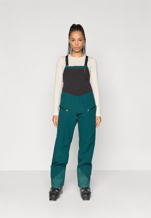 Tyrkysovo-čierne kombinézy s nastaviteľnými popruhmi, obsahujúce predné zipsové vrecká a zúžené nohy. Materiál vyzerá odolne s hladkým povrchom.