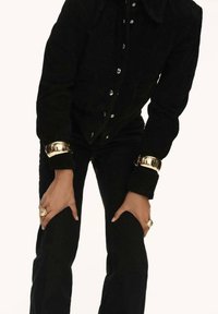 Giacca in suede nera con bottoni metallici, indossata con pantaloni neri aderenti. Manicotti e anelli dorati notevoli, che mettono in risalto un design elegante e moderno.