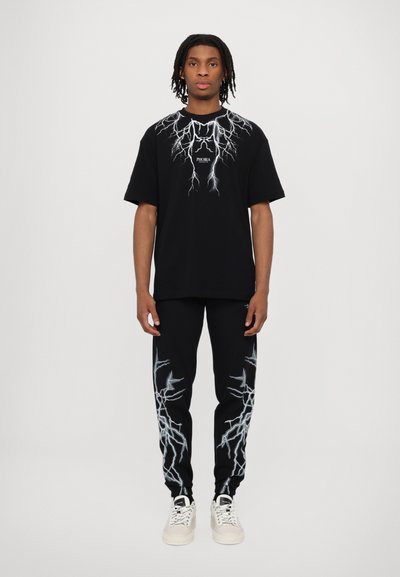 T-shirt nera in cotone con design fulmine sul petto, abbinata a pantaloni da tuta neri con accenti a forma di fulmine. Completata con sneaker bianche.