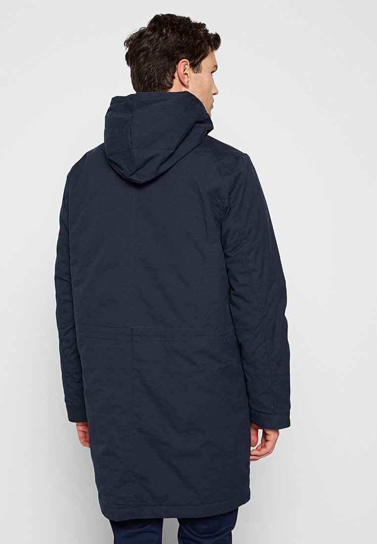 Parka bleu marine avec capuche, longueur longue, tissu lisse et deux poches latérales. Présente un design épuré et minimaliste.