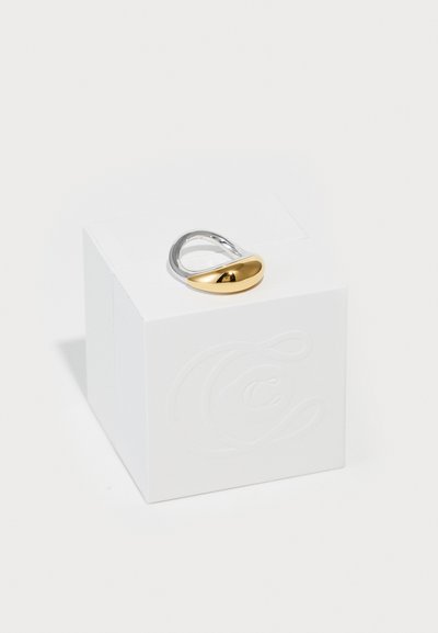 Zlatý a strieborný prsteň s zaobleným, lešteným dizajnom, položený na textúrovanej bielej krabičke. Prsteň sa vyznačuje bezšvovým prechodom farieb.