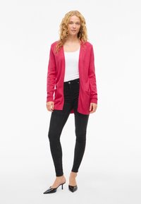 Long, open-front cardigan in bright pink knit fabric with ribbed cuffs, paired with black skinny jeans and black pointed-toe heels.