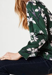 Grön blommig blus med vita och lila blommönster, långa ärmar, texturerad tyg, kombinerad med mörka jeans. Synlig hand som vilar på knäet.