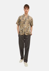 Chemise à manches courtes, brun à motifs avec des designs abstraits, associée à un pantalon sombre et des baskets blanches. Coupe décontractée avec un look relax.