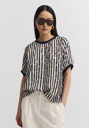 Frau mit langen dunklen Haaren, die große Sonnenbrille, goldene geometrische Ohrringe, ein gemustertes Kurzarmtop und weiße, hochtaillierte Hosen trägt.