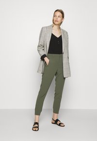 Blazer à quadros com punhos pretos, blusa preta com botões, calças verdes azeitona ajustadas e sandálias pretas. Design minimalista e texturas suaves.