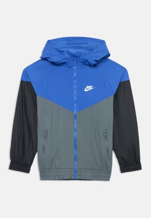 Veste coupe-vent Nike avec capuche, partie supérieure bleue, milieu gris et manches noires. Comprend deux poches zippées sur les côtés et des poignets élastiques.