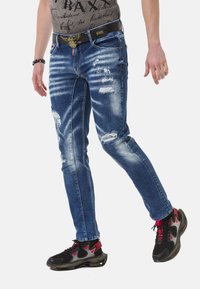 Blaue Jeans aus Denim mit schmaler Passform, die abgeriebene Stellen, Verwaschungen und einen schwarzen Gürtel mit einer dekorativen Schnalle aufweisen. Tragen Sie dazu schwarze und rote Sneaker.
