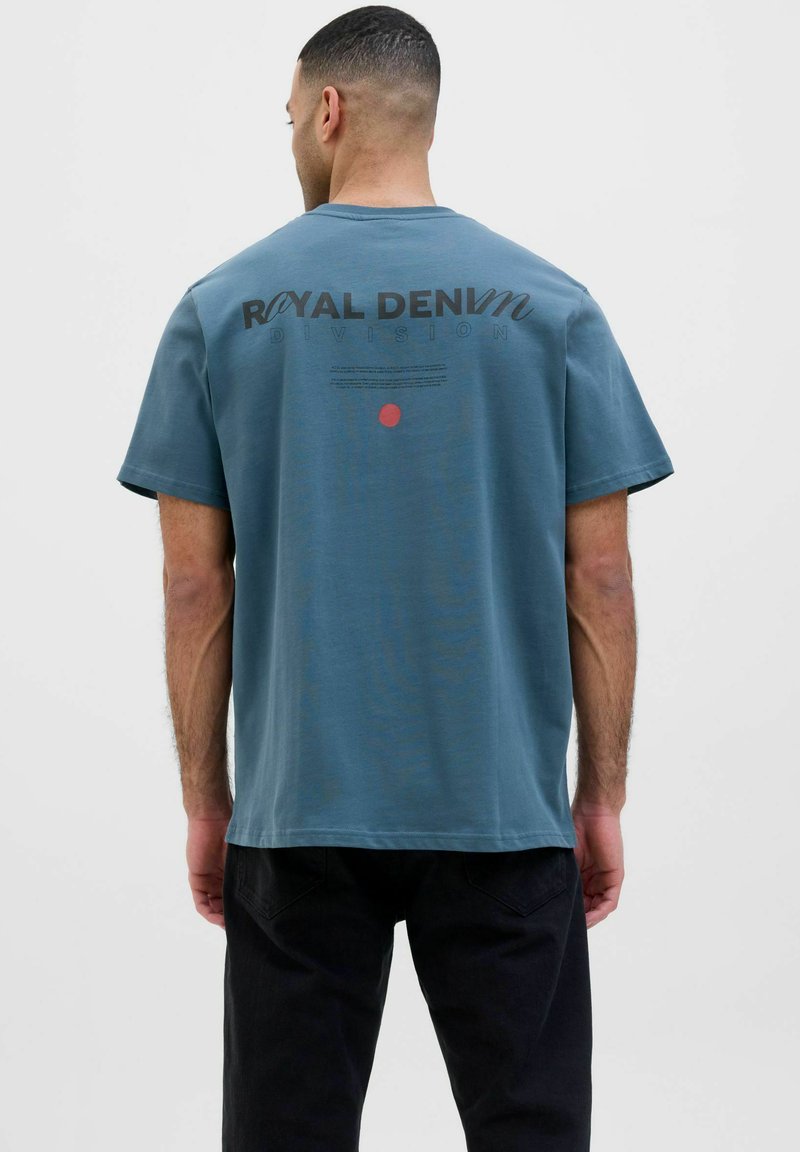 Zila kokvilnas T-kreklu ar īsām piedurknēm, kurā ir rakstīts "Royal Denim Division" un uz muguras ir sarkans aplis.
