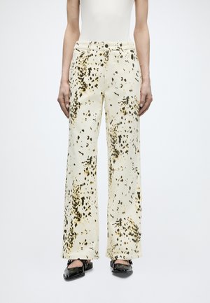 Cremefarbene gerade geschnittene Hose mit schwarzen und beigen abstrakten Flecken, getragen mit schwarzen Lackschuhen und einem weißen Oberteil, Frontalansicht von der Taille abwärts.