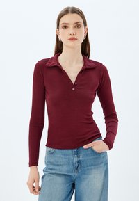 Burgundy pitkähihaisessa polo-paidassa on kaulus, nappilista ja pehmeä tekstuuri, joka on yhdistetty vaaleansinisiin farkkuihin. Yksinkertainen ja muutamaan muotoon istuva design.